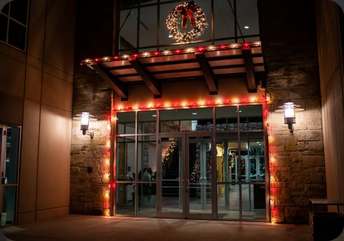 Commercial entrance outlined in red and warm white Christmas lights with a wreath above the doors
