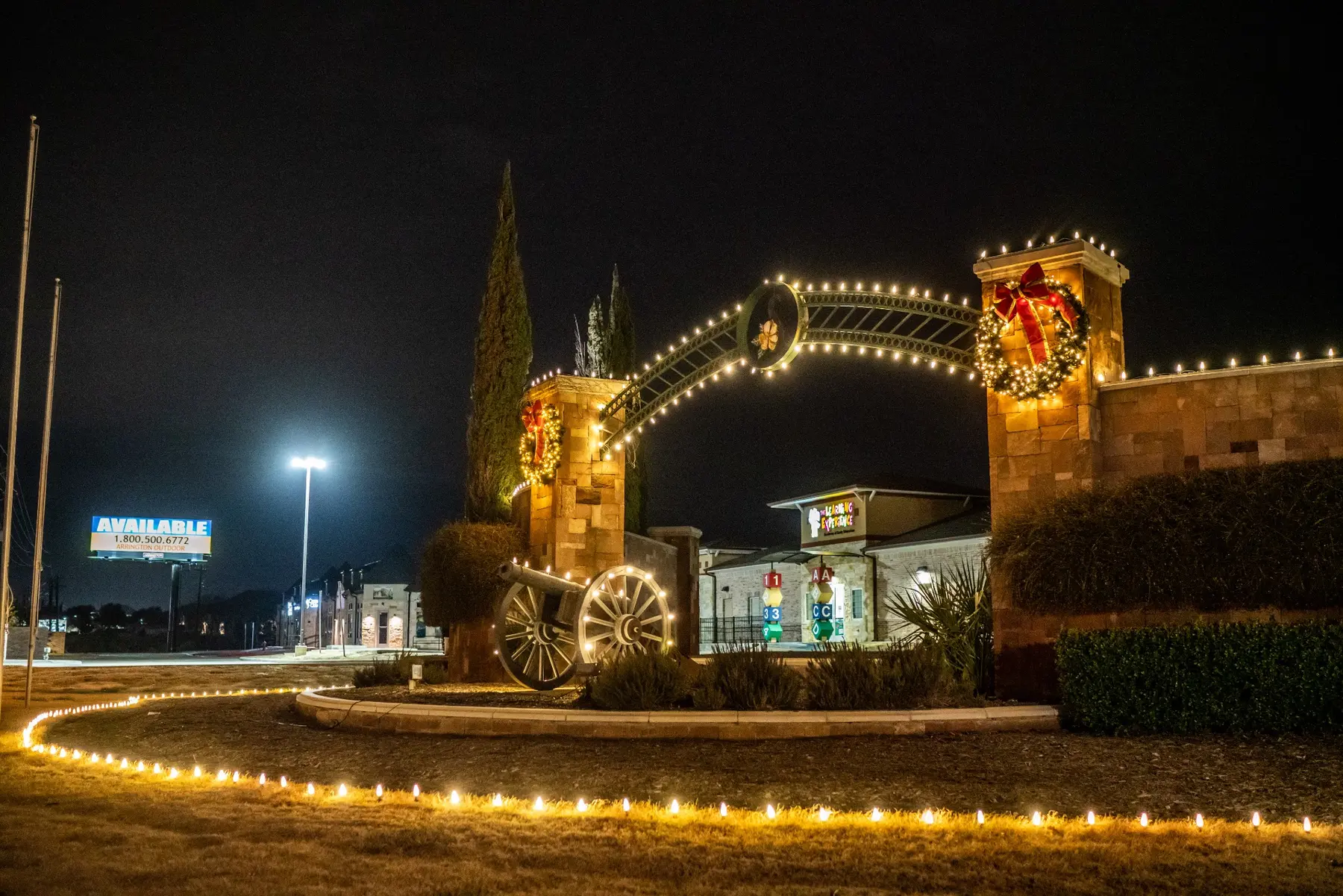 Stone community entrance with warm white stake lighting, a lit arch and holiday wreaths