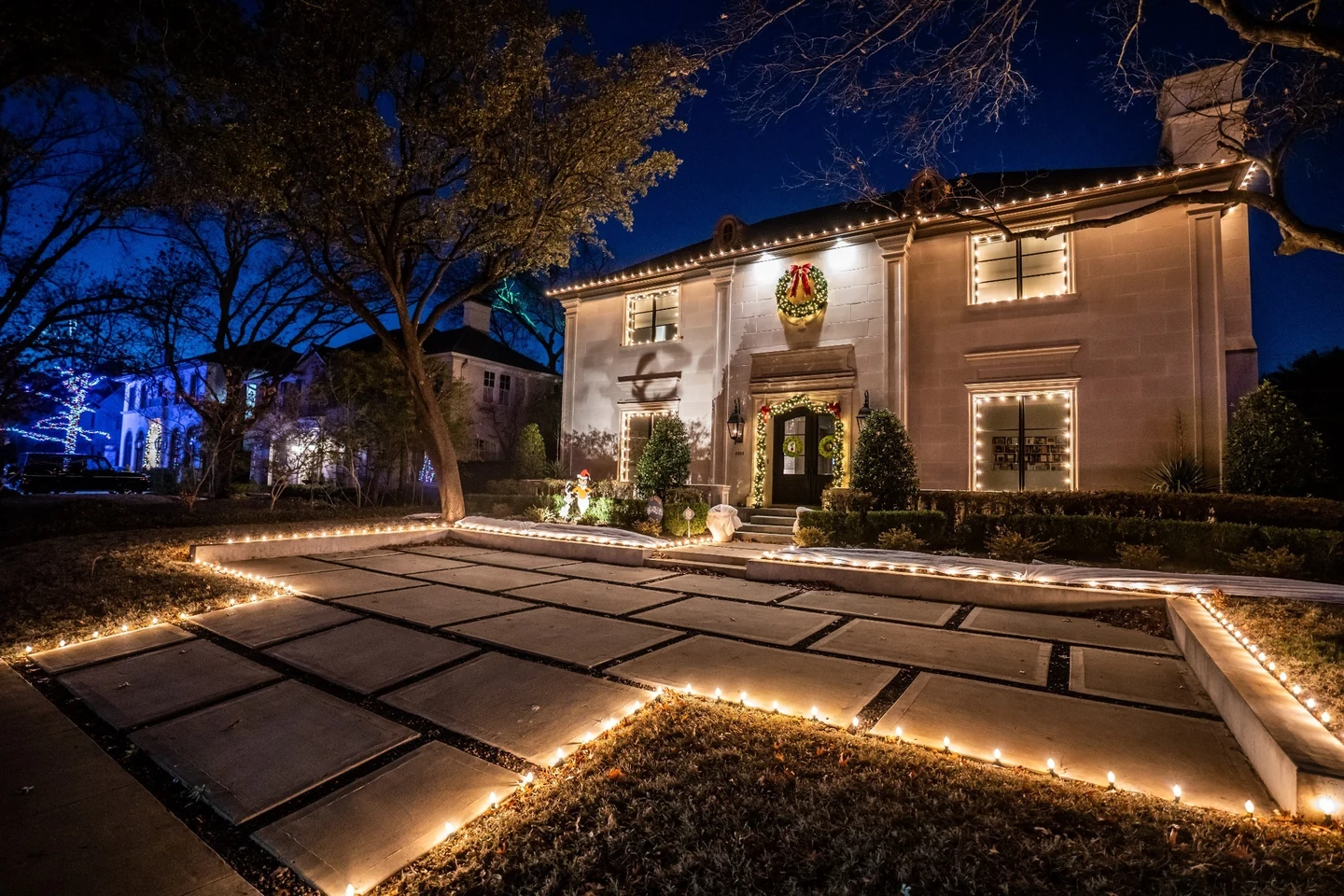 Two-story home with warm white roofline lighting, a lit wreath and landscape accents at night