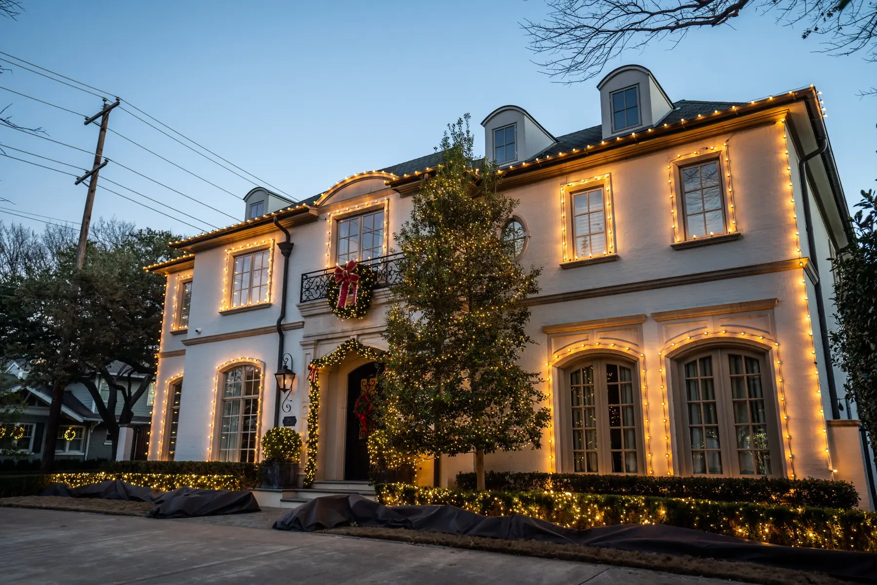 Two-story home with warm white roofline lights, wreaths and landscape accents at dusk
