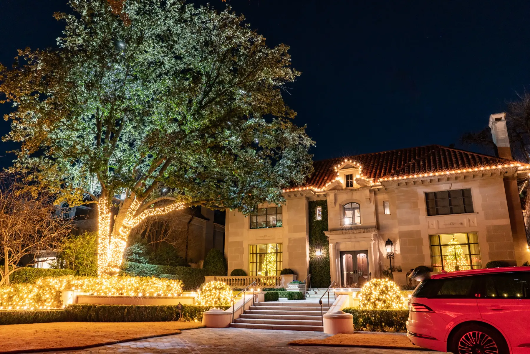 Large residence with a mature tree and landscaping illuminated in warm white holiday lights